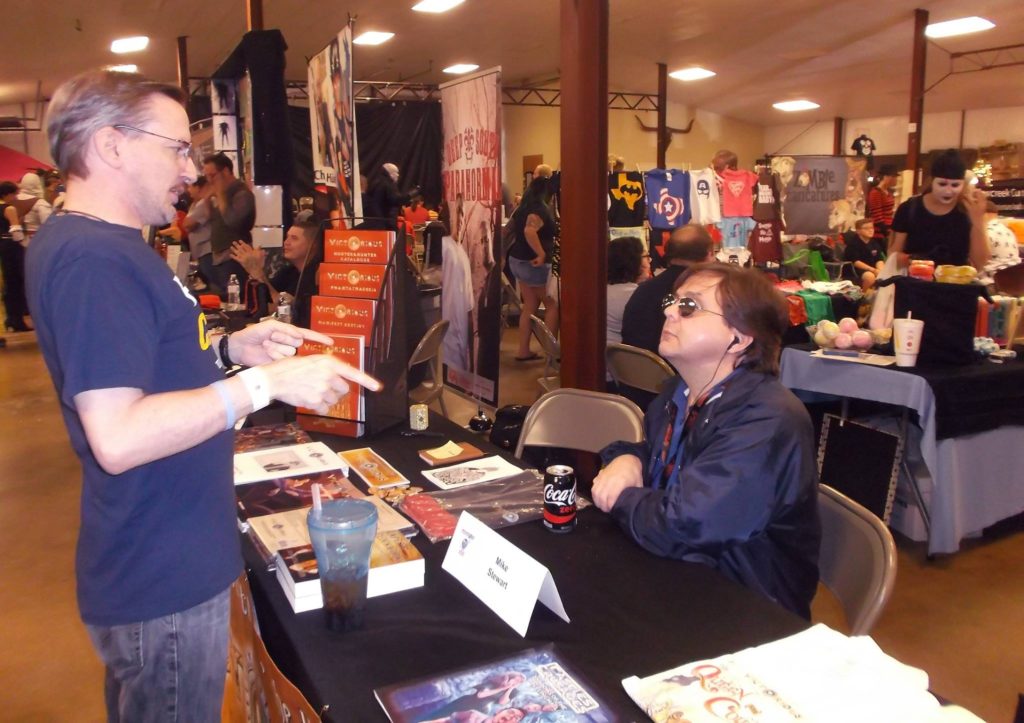 Author Mike Stewart sitting at table speaking with convention attendees.